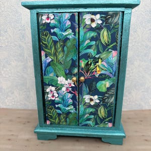 May include: A small, teal-painted cabinet with two doors. The doors feature a vibrant tropical floral print with white flowers and lush green leaves against a dark blue background. Gold-colored knobs complete the cabinet's design.