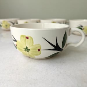 May include: A cream-colored ceramic teacup featuring a hand-painted design of a pale yellow four-petaled flower with small red dots on the petal tips, accompanied by dark green leaves and black stems, with a matching handle on the right side.