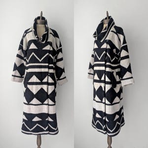 May include: A long, black, and cream-colored geometric patterned coat. The coat features a shawl collar, a single button closure, and two pockets. The coat is displayed on a mannequin.