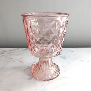 May include: A light pink glass goblet featuring a textured diamond pattern on the bowl and a vertical ribbed design on the pedestal base. The vessel has a wide, circular rim and a clear, translucent finish that catches the light.
