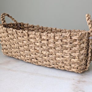 May include: A rectangular woven basket with two braided handles. The basket is made of natural-colored fibers, with a tightly woven pattern. The basket is sitting on a white marble surface.