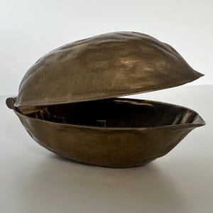 May include: A bronze-colored, open clam shell-shaped container. The top shell is lifted, revealing the interior. The container has a textured surface and is set against a white background. The object appears to be made of metal.