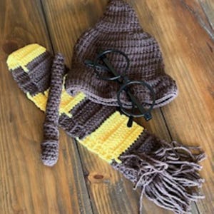 May include: A brown crocheted pointed hat, a brown and yellow striped crocheted scarf with fringe, a brown crocheted wand, and a pair of black round-rimmed glasses. The items are arranged together to form a set of accessories with a consistent color scheme.