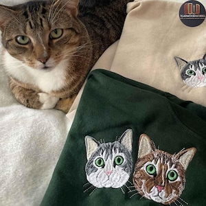 Può includere: Un gatto tigrato riposa vicino a due magliette ricamate. Una maglietta è verde scuro, che mostra due facce di gatto con occhi verdi. L'altra maglietta è beige, con una singola faccia di gatto. Le magliette sono ricamate con facce di gatto dettagliate.