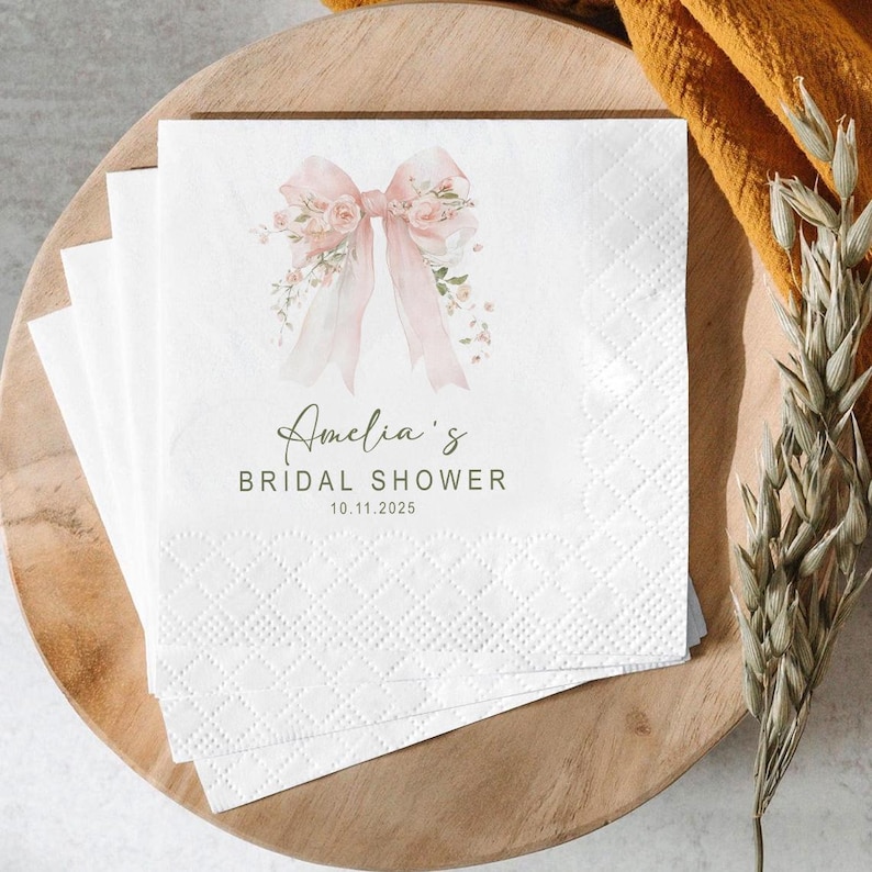 May include: White paper napkins with a pink bow and floral design, with the text "Amelia's Bridal Shower 10.11.2025". The napkins are stacked on a wooden tray, with a yellow cloth and dried plants in the background.
