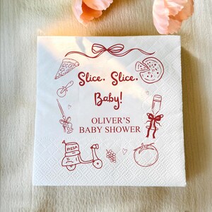 Op de afbeelding: Witte papieren servet met rode illustraties van pizzapunten, een pizzasnijder, een scooter en andere pizza-thema graphics. De tekst "Slice. Slice. Baby!" en "Oliver's Baby Shower" zijn rood gedrukt.