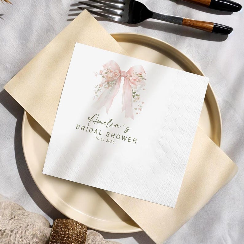 May include: A square white napkin with a watercolor floral bow design and the text "Amelia's BRIDAL SHOWER 10.11.2025". The napkin is placed on a beige plate with a folded beige napkin and a fork.