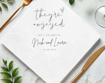 May include: White square napkin with the text "they're engaged" in elegant script. Below, it reads "Let's Celebrate Nick and Laura" with the date 10.08.2025. Gold fork and knife are on the sides, with greenery accents, suggesting a wedding or engagement theme.