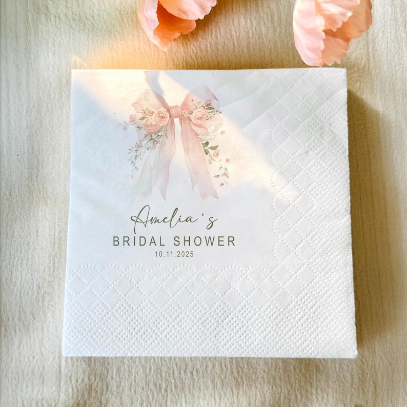 May include: White paper napkin with a floral and ribbon design. The napkin is printed with the text "Amelia's Bridal Shower" and the date 10.11.2025. The napkin has a quilted pattern.