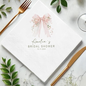 May include: White square paper napkin with a watercolor design of a pink bow and floral accents. The text "Amelia's Bridal Shower 10.11.2025" is printed in a script font. Gold-colored fork and knife are visible.