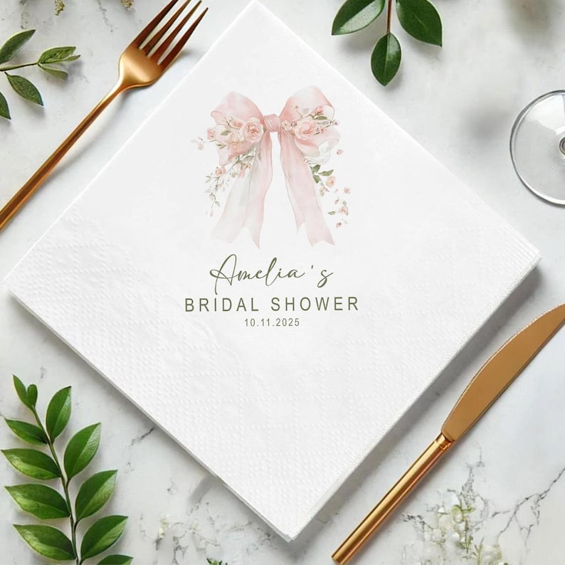 May include: White square paper napkin with a watercolor design of a pink bow and floral accents. The text "Amelia's Bridal Shower 10.11.2025" is printed in a script font. Gold-colored fork and knife are visible.