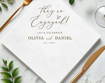May include: White square paper napkin with the text "They're Engaged!" in brown script. Below the text are the words "LET'S CELEBRATE" and the names "OLIVIA and DANIEL" with the date "20.11.2025". A gold fork and knife are on the sides.