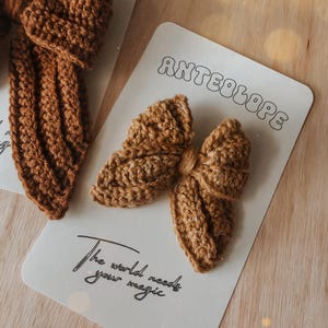 Puede incluir: Primer plano de lazos para el pelo de ganchillo marr&oacute;n hechos a mano sobre tarjetas blancas. Las tarjetas tienen la palabra "ANTEOLOPE" en una fuente retro y la frase "The world needs your magic" en cursiva. Los lazos est&aacute;n sobre una superficie de madera clara.