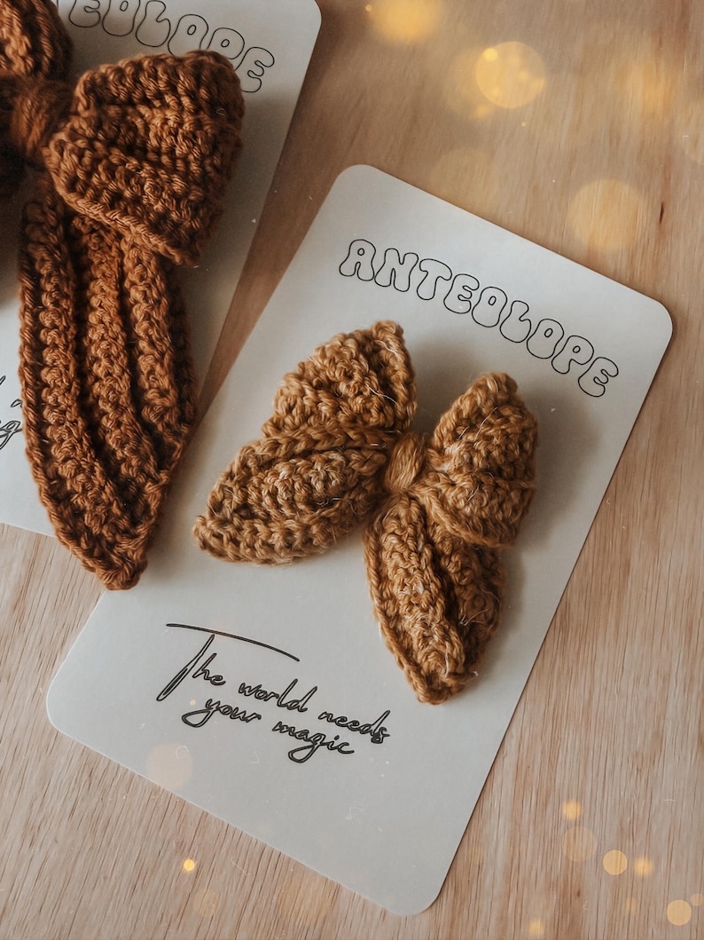 Puede incluir: Primer plano de lazos para el pelo de ganchillo marr&oacute;n hechos a mano sobre tarjetas blancas. Las tarjetas tienen la palabra "ANTEOLOPE" en una fuente retro y la frase "The world needs your magic" en cursiva. Los lazos est&aacute;n sobre una superficie de madera clara.