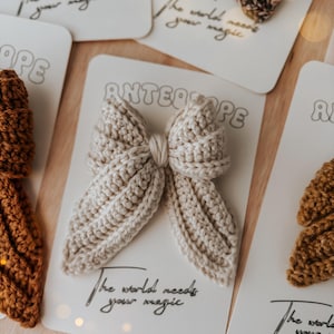 Puede incluir: Una colecci&oacute;n de lazos para el cabello hechos a mano con ganchillo en varios colores, incluyendo beige y marr&oacute;n. Cada lazo se muestra en una tarjeta blanca con el texto "The world needs your magic" y el nombre de la marca "ANTEAOPE".