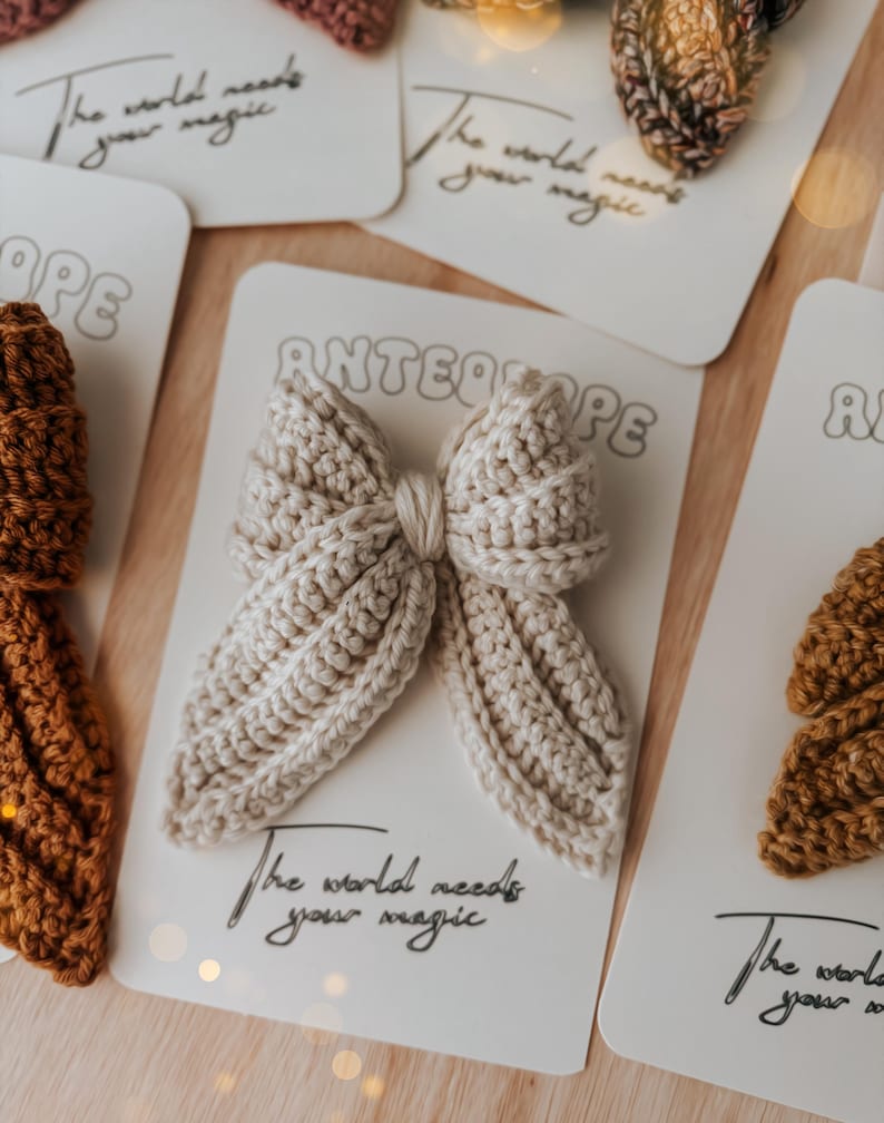 Puede incluir: Una colecci&oacute;n de lazos para el cabello hechos a mano con ganchillo en varios colores, incluyendo beige y marr&oacute;n. Cada lazo se muestra en una tarjeta blanca con el texto "The world needs your magic" y el nombre de la marca "ANTEAOPE".