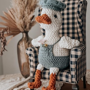 May include: A handmade crochet goose doll wearing a blue beret and overalls, seated in a plaid armchair. The goose has a white body, orange beak and feet, and small flower accents. The chair has a black and white checkered pattern.