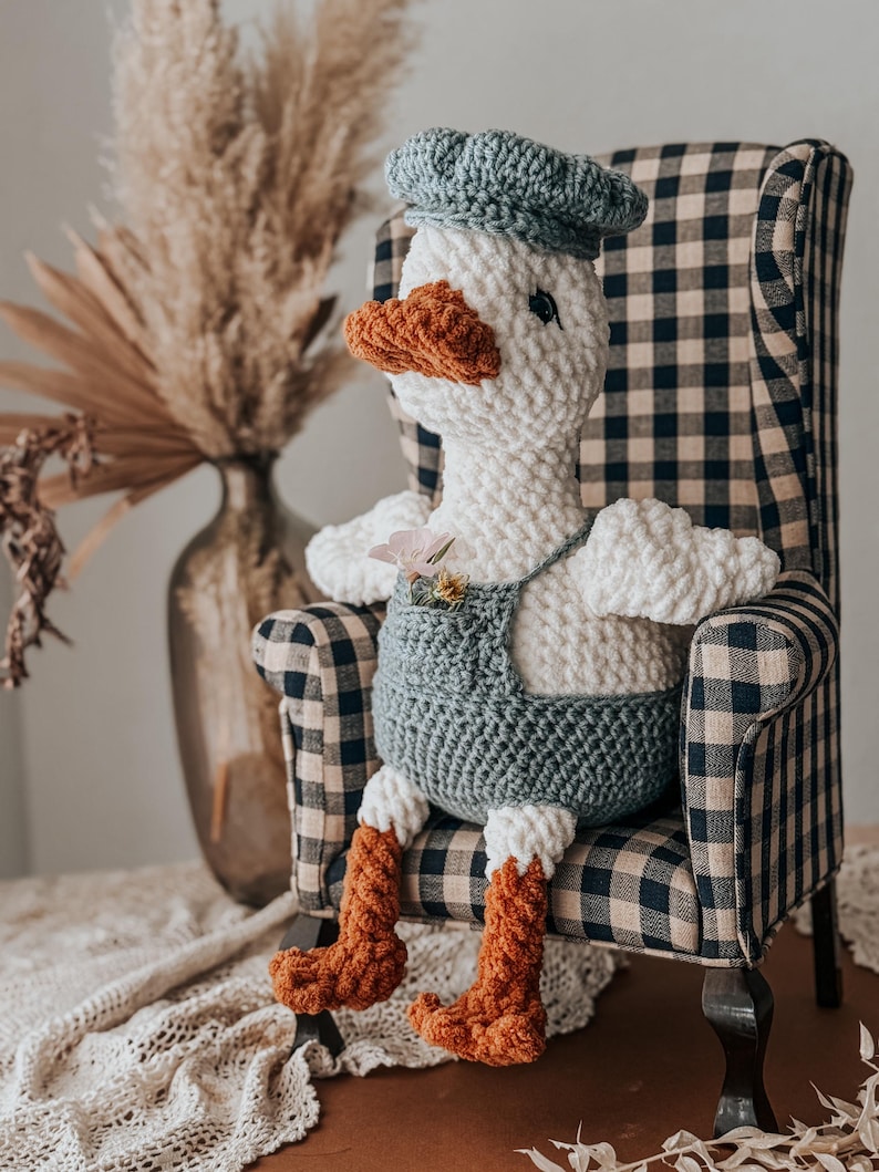 May include: A handmade crochet goose doll wearing a blue beret and overalls, seated in a plaid armchair. The goose has a white body, orange beak and feet, and small flower accents. The chair has a black and white checkered pattern.