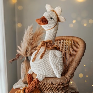 May include: A handmade, white crochet goose doll with a long neck, orange beak, and feet. The goose is seated in a wicker chair and has a flower-like headpiece. The doll has brown accents and is a soft, plush toy.