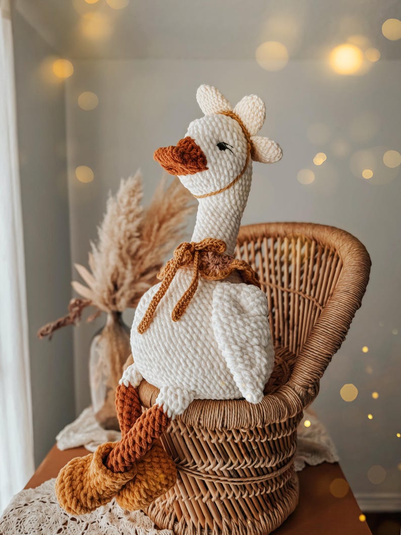 May include: A handmade, white crochet goose doll with a long neck, orange beak, and feet. The goose is seated in a wicker chair and has a flower-like headpiece. The doll has brown accents and is a soft, plush toy.