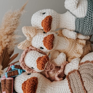 May include: A stack of handmade crochet goose plushies. The geese are primarily white with brown beaks and accents. Some geese wear pastel-colored clothing. The plushies are arranged on a woven surface, with spools of thread visible.