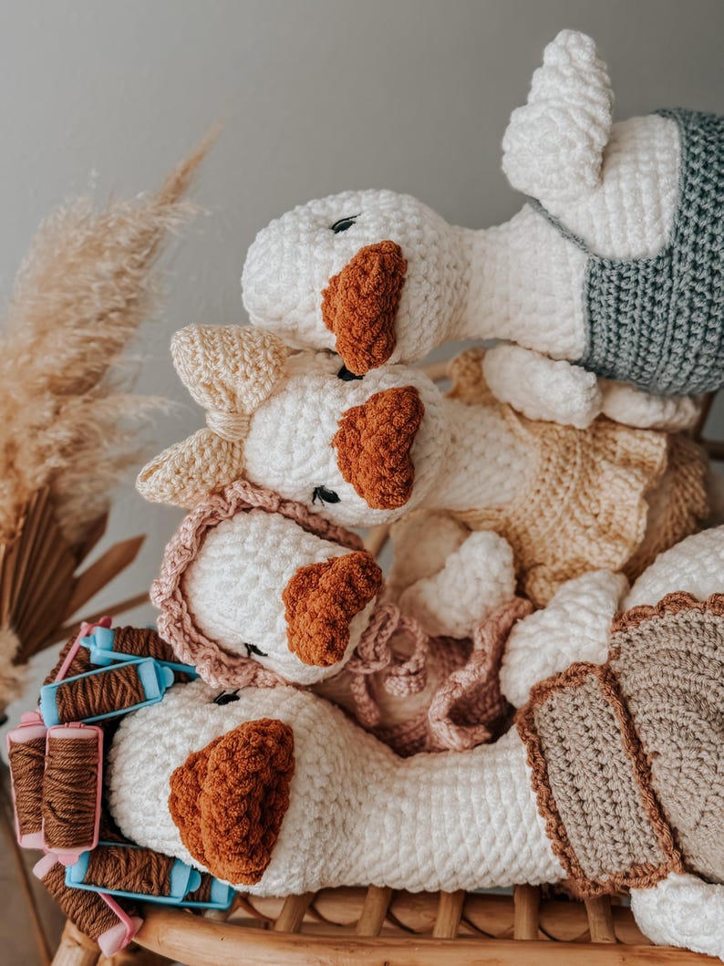 May include: A stack of handmade crochet goose plushies. The geese are primarily white with brown beaks and accents. Some geese wear pastel-colored clothing. The plushies are arranged on a woven surface, with spools of thread visible.