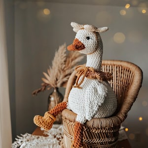 May include: A handmade crochet goose doll with a white body, brown beak, and orange legs and feet. The goose wears a matching hat and a decorative collar. It is seated in a wicker chair, creating a cozy, rustic aesthetic.