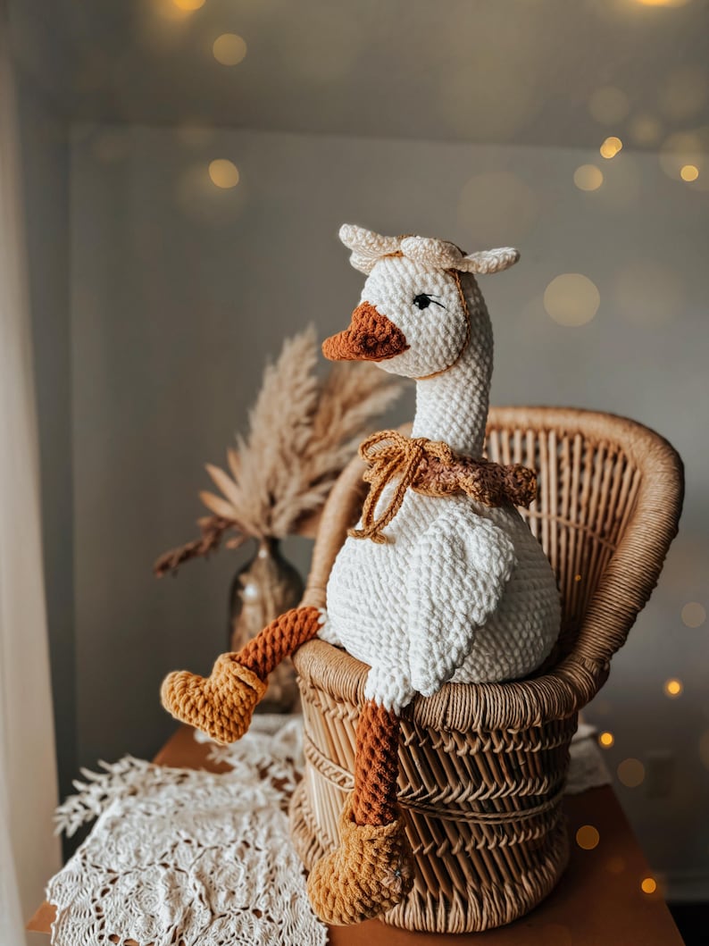 May include: A handmade crochet goose doll with a white body, brown beak, and orange legs and feet. The goose wears a matching hat and a decorative collar. It is seated in a wicker chair, creating a cozy, rustic aesthetic.