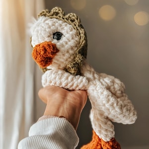 Pu&ograve; includere: Un giocattolo di peluche all'uncinetto a forma di oca con corpo bianco, zampe marroni e testa verde. L'oca &egrave; tenuta sollevata da una mano.