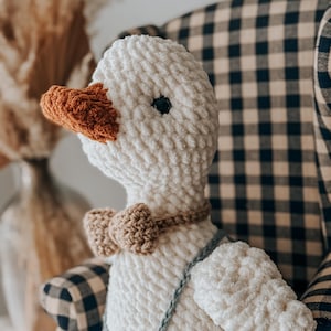 May include: A handmade white crochet goose doll with an orange beak and a brown bow tie. The goose wears a blue crocheted overall. The doll is sitting in a checkered armchair. A decorative item for a nursery or child's room.