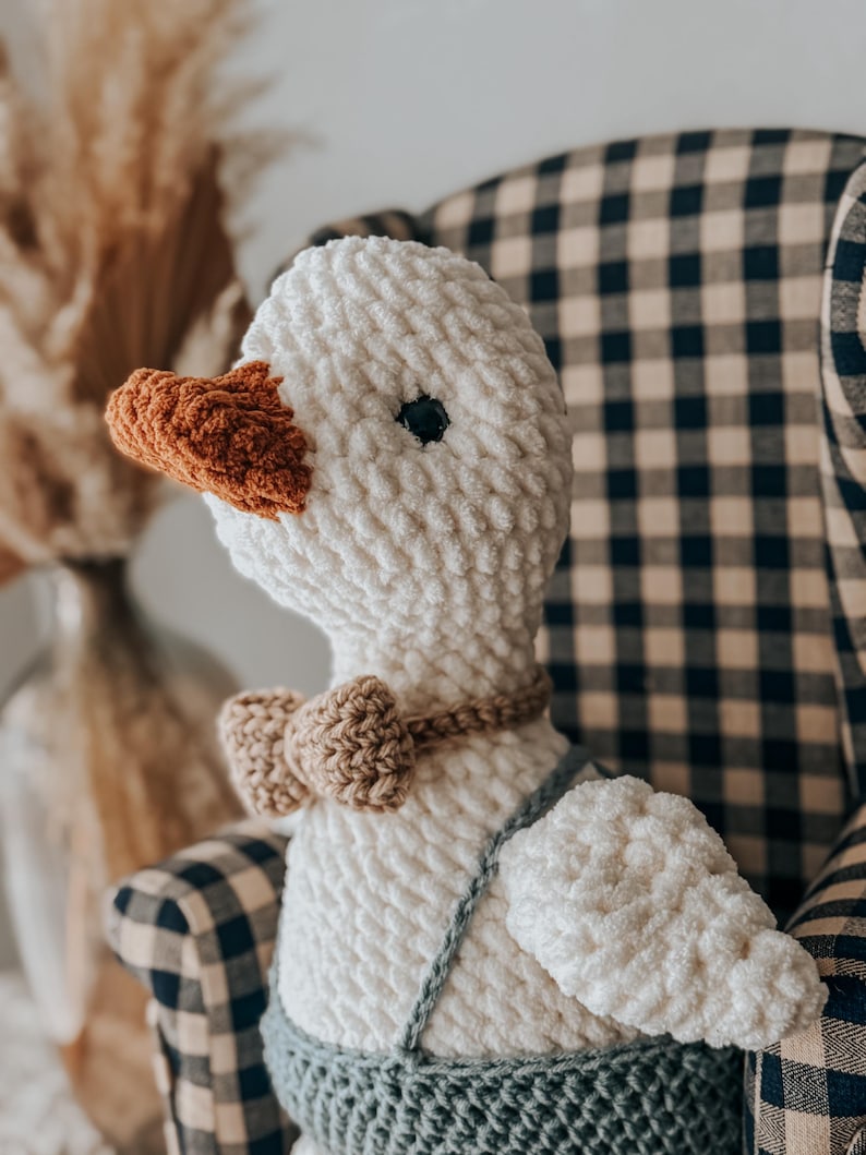 May include: A handmade white crochet goose doll with an orange beak and a brown bow tie. The goose wears a blue crocheted overall. The doll is sitting in a checkered armchair. A decorative item for a nursery or child's room.