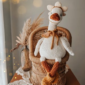 May include: A handmade, white crochet goose doll with a flower crown and a brown beak and feet. The goose is seated in a small wicker chair. The doll has a brown collar and is a whimsical decorative item.