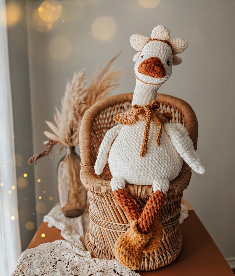 May include: A handmade, white crochet goose doll with a flower crown and a brown beak and feet. The goose is seated in a small wicker chair. The doll has a brown collar and is a whimsical decorative item.