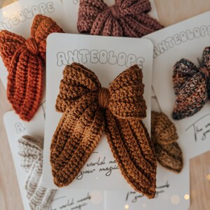 Puede incluir: Colecci&oacute;n de lazos para el cabello hechos a mano con ganchillo en varios colores, incluyendo &oacute;xido, marr&oacute;n y malva. Cada lazo est&aacute; unido a una tarjeta blanca con la palabra "ANTEOLOPE". Los lazos est&aacute;n dispuestos sobre una superficie de madera clara.
