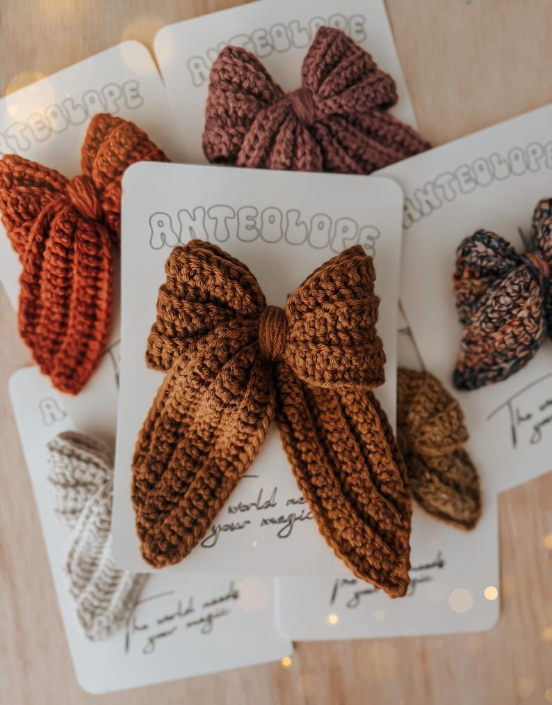 Puede incluir: Colecci&oacute;n de lazos para el cabello hechos a mano con ganchillo en varios colores, incluyendo &oacute;xido, marr&oacute;n y malva. Cada lazo est&aacute; unido a una tarjeta blanca con la palabra "ANTEOLOPE". Los lazos est&aacute;n dispuestos sobre una superficie de madera clara.