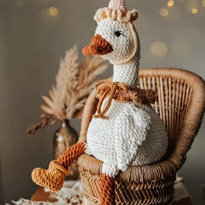 May include: A handmade, white crochet goose doll with a brown beak, feet, and a decorative neck piece. The goose wears a pink and white party hat and sits in a wicker chair. The doll is a whimsical decorative item.