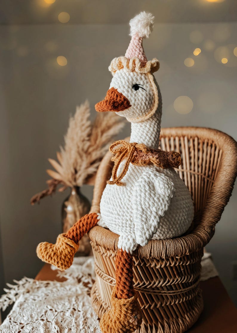 May include: A handmade, white crochet goose doll with a brown beak, feet, and a decorative neck piece. The goose wears a pink and white party hat and sits in a wicker chair. The doll is a whimsical decorative item.
