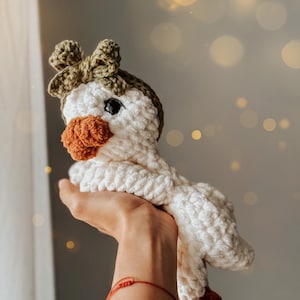 Pu&ograve; includere: Un giocattolo di oca in crochet bianco con un becco marrone e un fiocco verde. L'oca &egrave; tenuta da una mano con un braccialetto rosso.