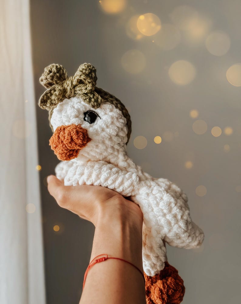 Pu&ograve; includere: Un giocattolo di oca in crochet bianco con un becco marrone e un fiocco verde. L'oca &egrave; tenuta da una mano con un braccialetto rosso.