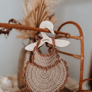 May include: A handmade, crochet baby bib in a neutral beige color with a scalloped brown border. The bib hangs on a wooden hanger shaped like a swan. The word "ASTROLOPE" is etched on the hanger.