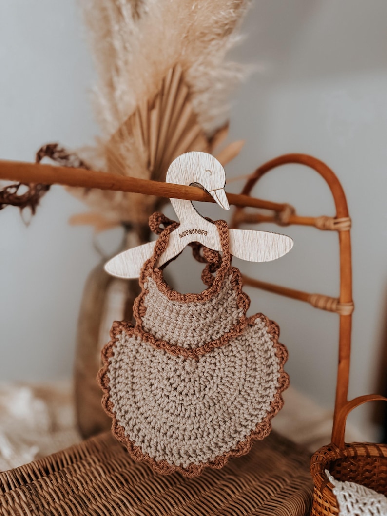 May include: A handmade, crochet baby bib in a neutral beige color with a scalloped brown border. The bib hangs on a wooden hanger shaped like a swan. The word "ASTROLOPE" is etched on the hanger.
