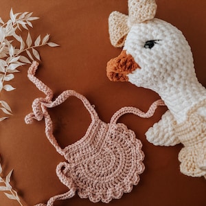 May include: A crochet goose doll with a bow and a pink crochet bib on a brown surface. The goose has a white body, a beige dress, and an orange beak and feet. A sprig of white leaves is on the left.