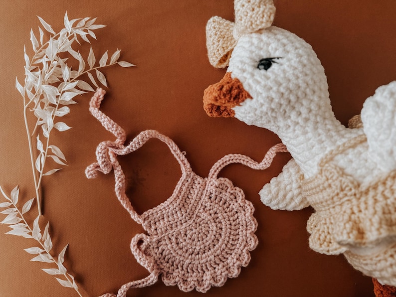 May include: A crochet goose doll with a bow and a pink crochet bib on a brown surface. The goose has a white body, a beige dress, and an orange beak and feet. A sprig of white leaves is on the left.