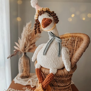 May include: A handmade, white crochet goose doll with a party hat, scarf, and boots. The goose is seated in a wicker chair. The hat is pink and yellow, the scarf is gray, and the boots are orange and yellow.