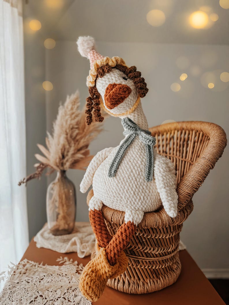 May include: A handmade, white crochet goose doll with a party hat, scarf, and boots. The goose is seated in a wicker chair. The hat is pink and yellow, the scarf is gray, and the boots are orange and yellow.