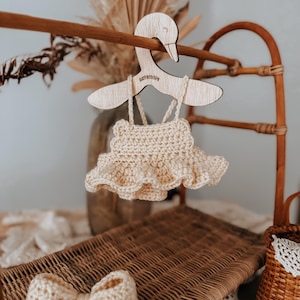 May include: A cream-colored, crocheted baby dress with a ruffled skirt hangs from a wooden swan-shaped hanger. A matching crocheted bow is on a wicker surface. The hanger has the text "BABY SCOOP" engraved on it.