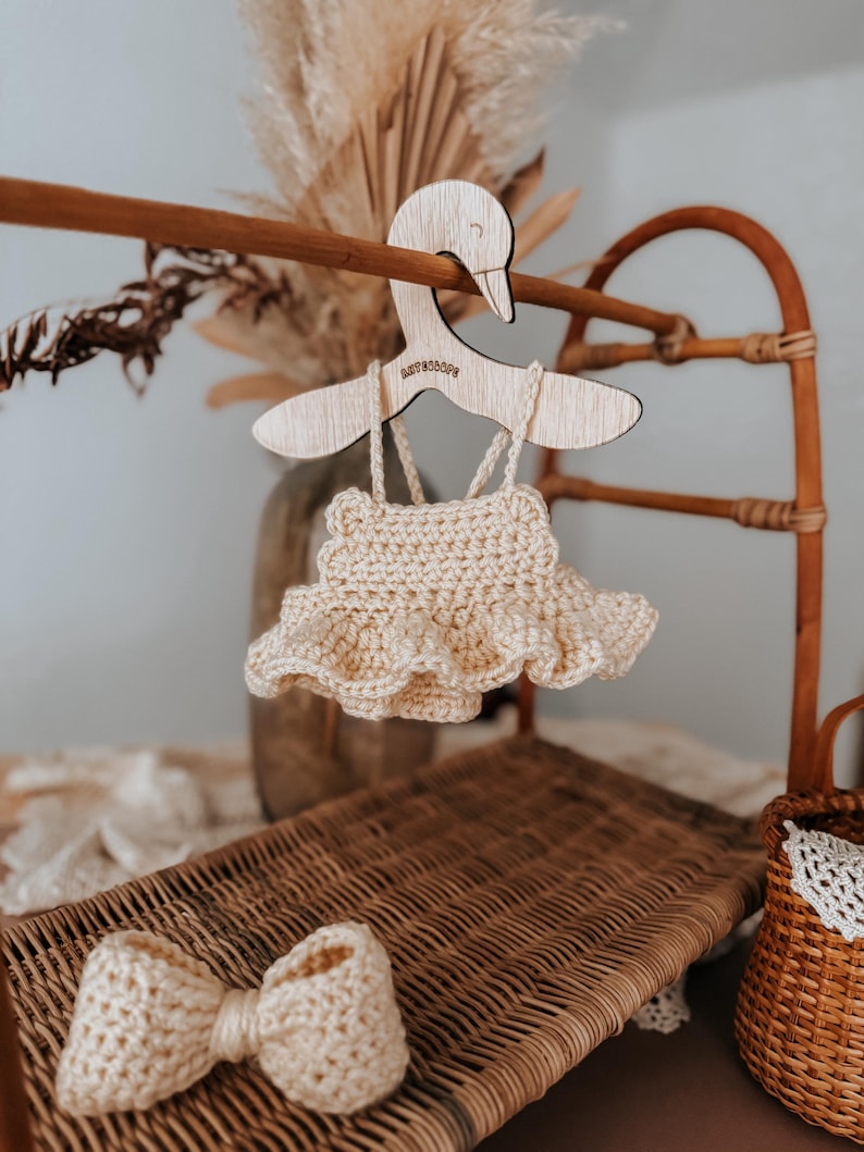 May include: A cream-colored, crocheted baby dress with a ruffled skirt hangs from a wooden swan-shaped hanger. A matching crocheted bow is on a wicker surface. The hanger has the text "BABY SCOOP" engraved on it.