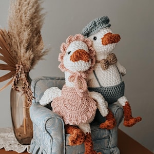 May include: Two handmade crochet geese dolls, one wearing a pink dress and bonnet, the other a blue cap and suspenders. They are seated on a small, patterned blue armchair, with orange feet and beaks.