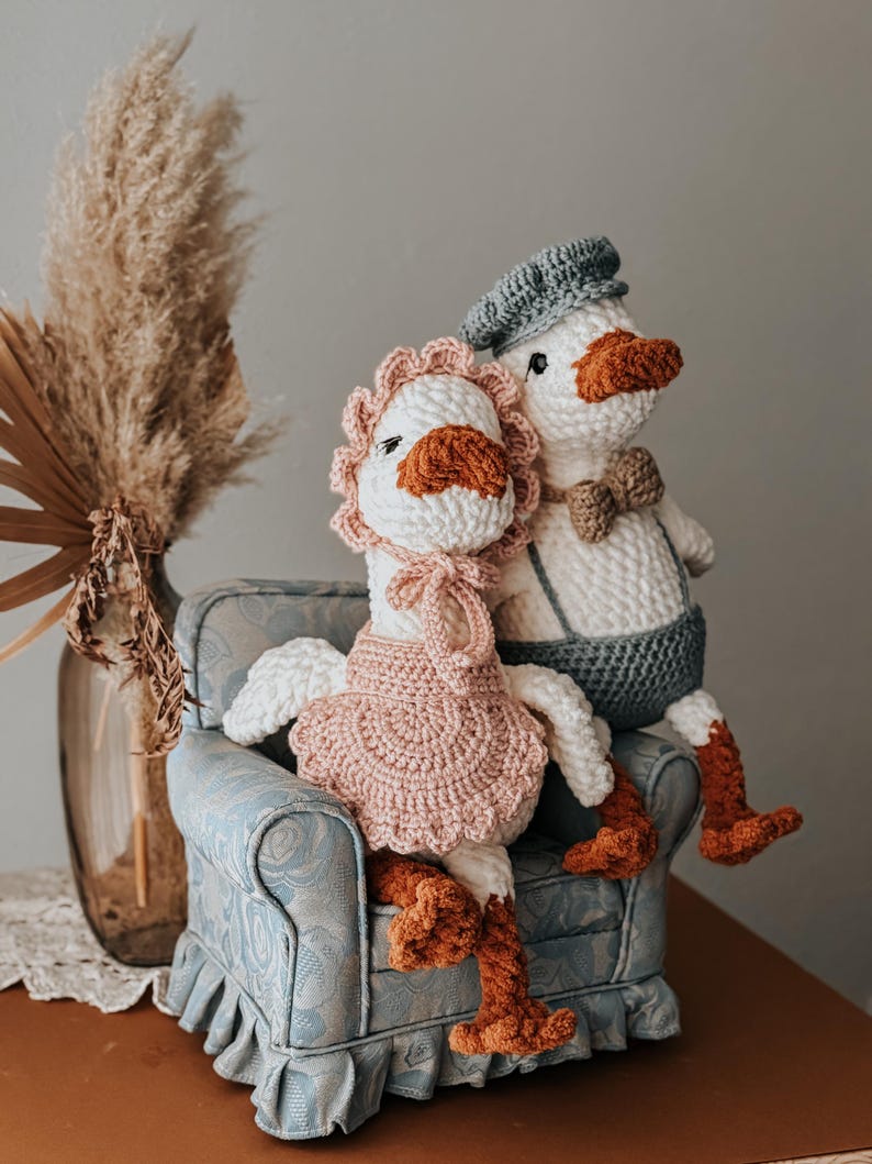 May include: Two handmade crochet geese dolls, one wearing a pink dress and bonnet, the other a blue cap and suspenders. They are seated on a small, patterned blue armchair, with orange feet and beaks.