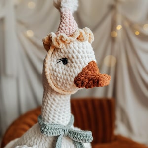 May include: A handmade, white crochet goose wearing a party hat with a pink and white gradient and a light green bow tie. The goose has an orange beak and black eyes. The plush toy is designed for children.